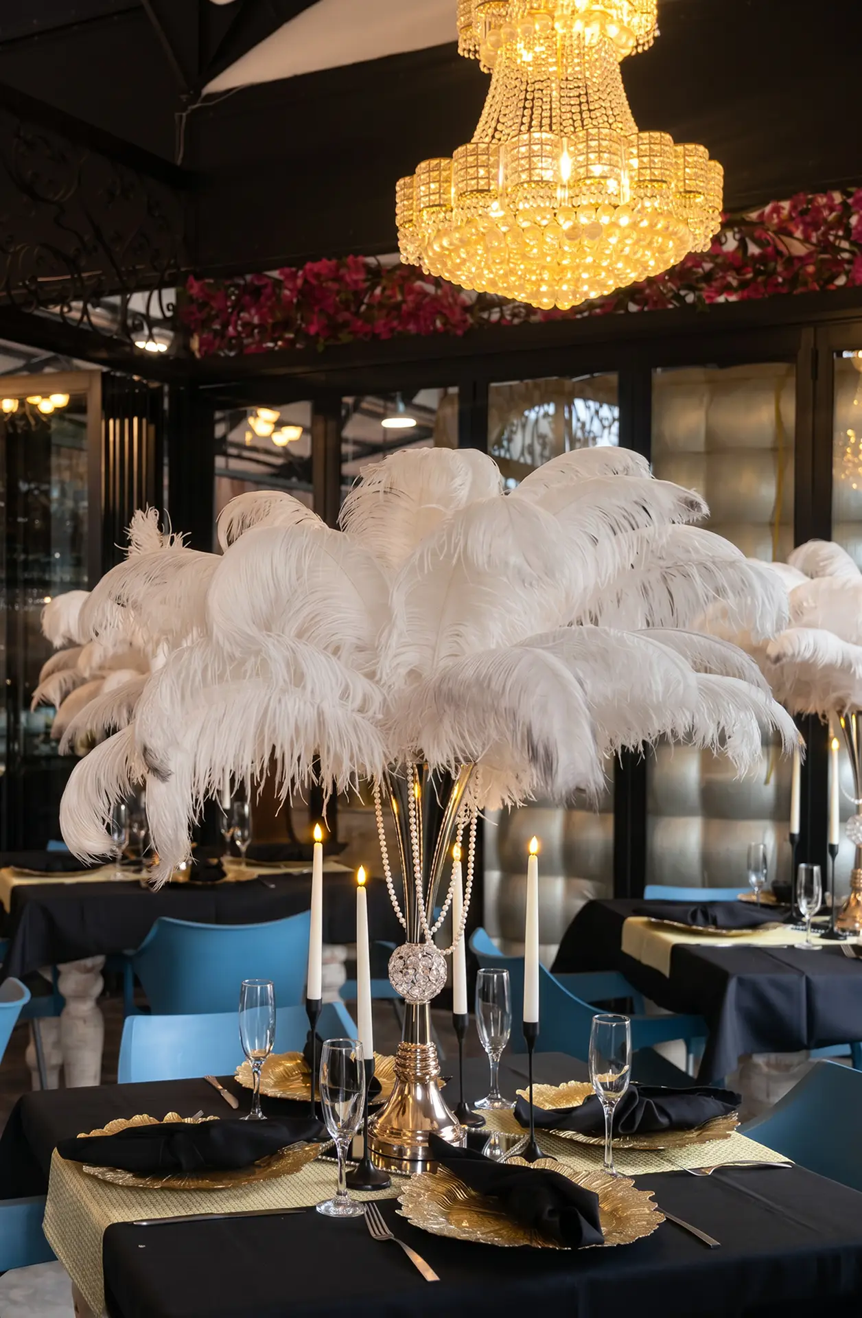 Elegant dining table with gold plates, black napkins, white feather centerpiece, candles, and blue chairs.