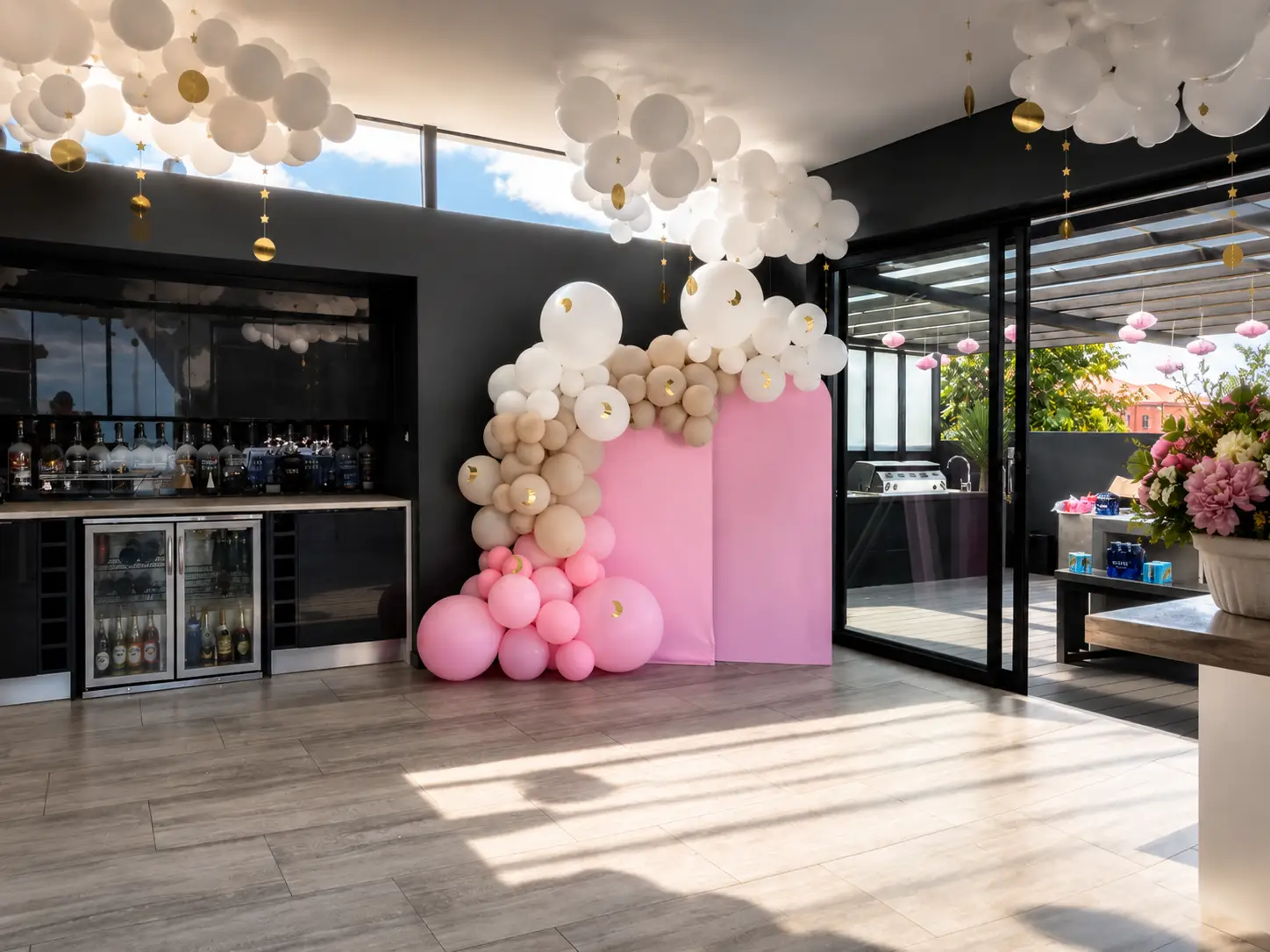 Modern indoor party setup with pink, beige, and white balloons and a mini bar area with drinks.