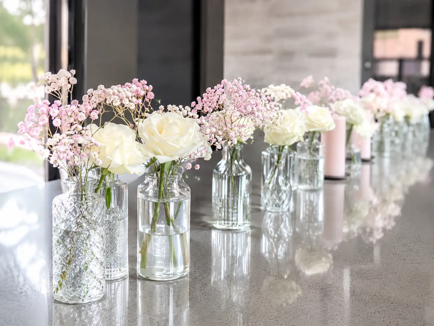 Row of glass vases with white roses and small pink flowers on a polished stone countertop.