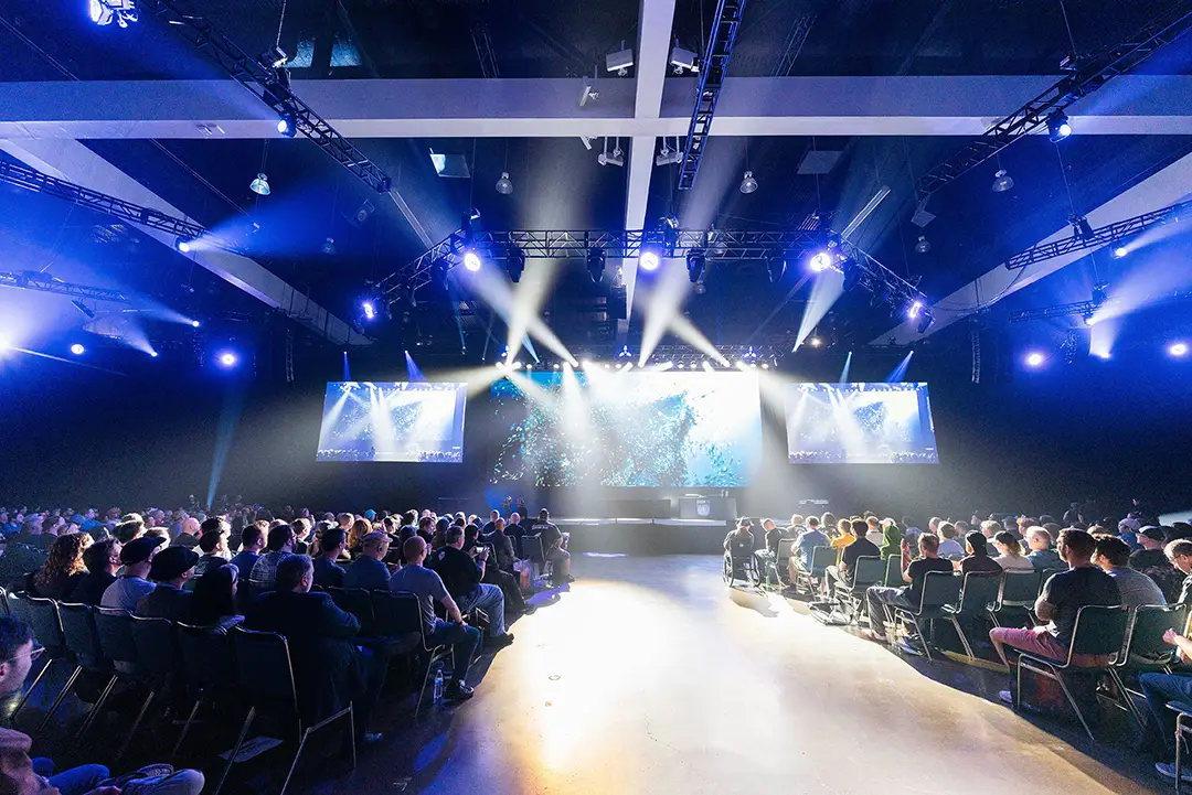 Crowded audience seated facing a stage with large screens and bright spotlights in a dark auditorium.