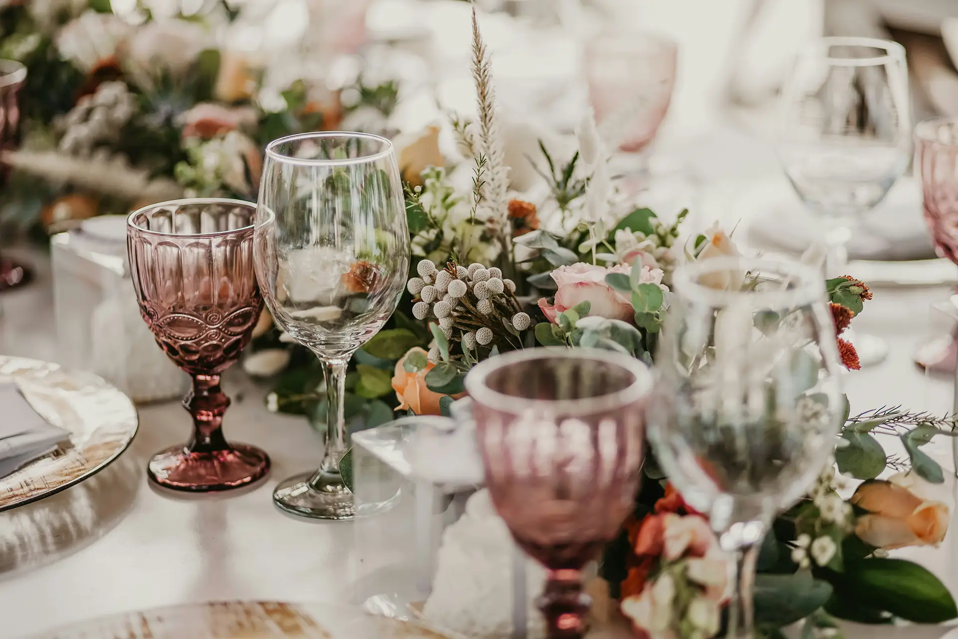 Elegant table setting with clear and purple wine glasses and a floral centerpiece with roses and greenery.