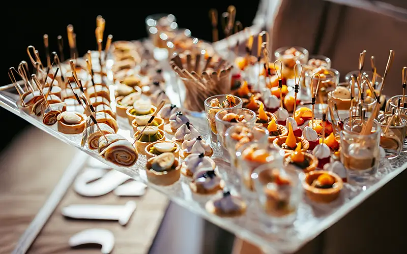 Tray of assorted elegant bite-sized appetizers and desserts on skewers and in small glasses.