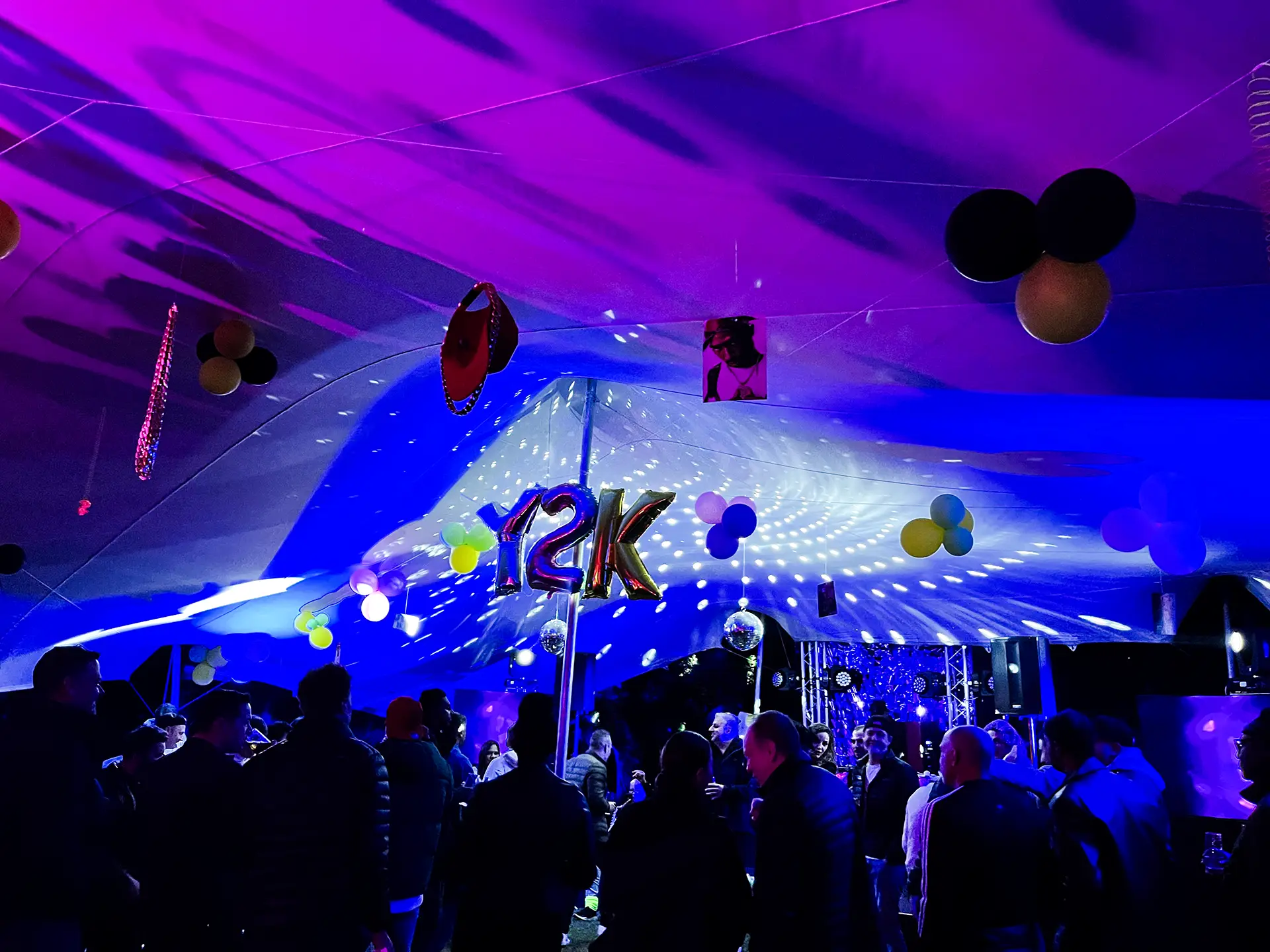 People gathered under a tent with blue and purple lighting and Y2K balloons and decorations.