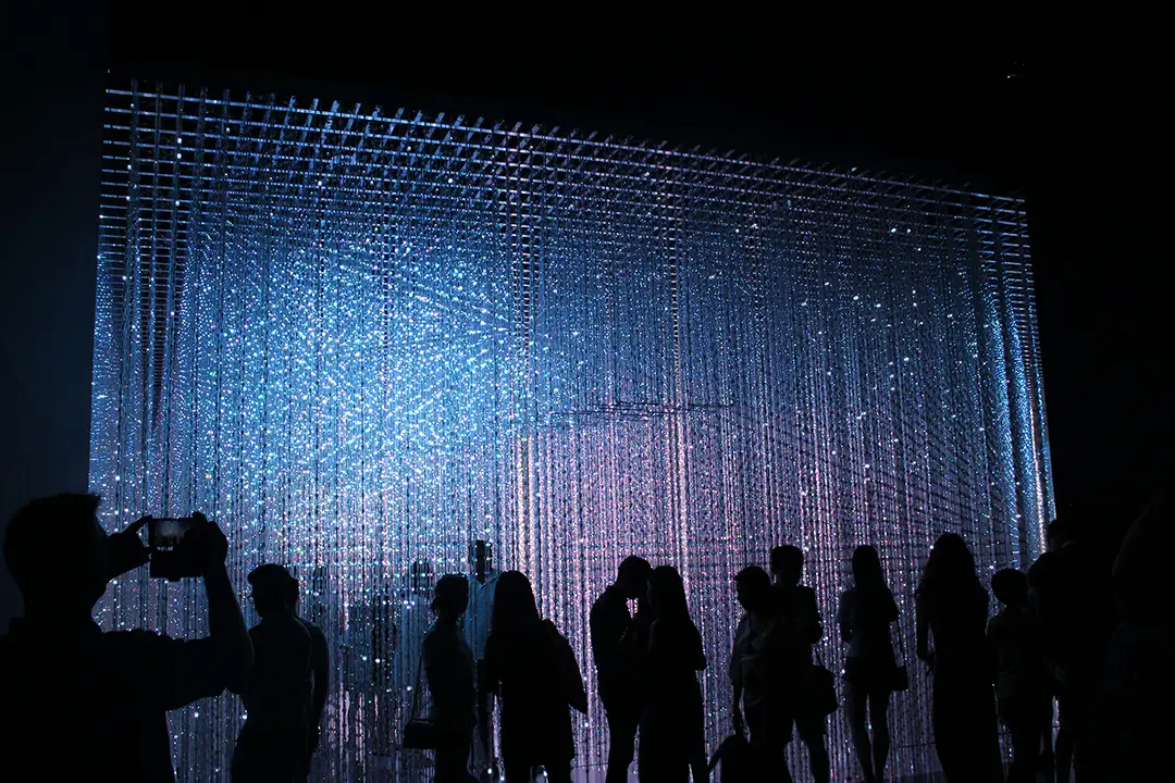 Silhouettes of people in front of a large illuminated art installation with hanging lights.