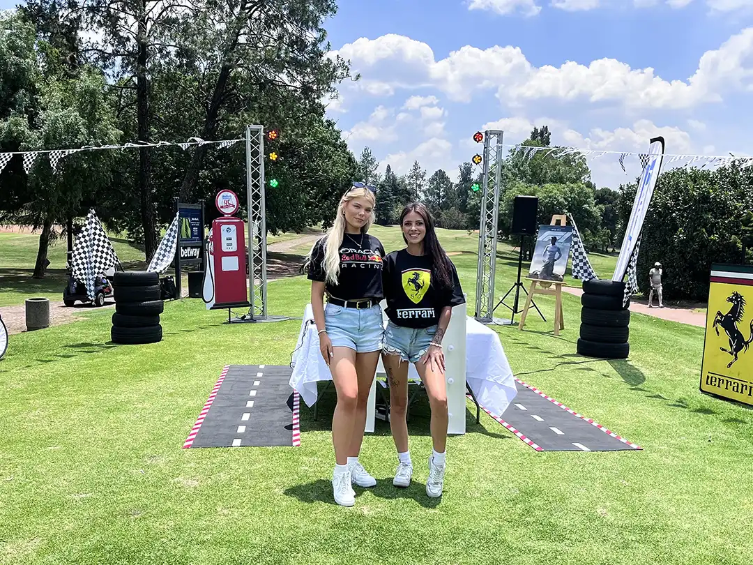 Two young women wearing Formula 1 shirts standing on grass with race-themed decorations behind them.