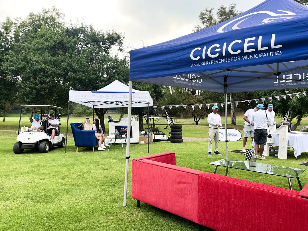 Golf event tents with people chatting, golf carts, couches, and checkered flags on a green course.