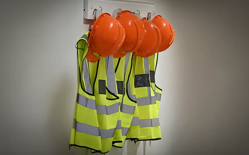 Four orange hard hats hanging on a wall rack above four yellow reflective safety vests.