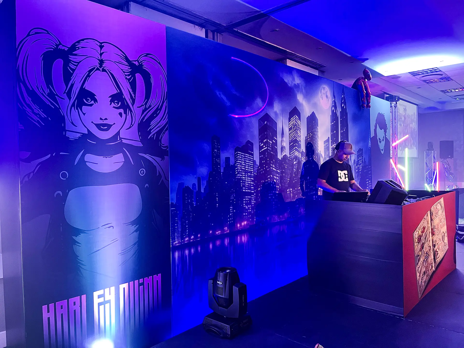 DJ wearing DC shirt performs beside comic-style murals of a city skyline and female character with pigtails.