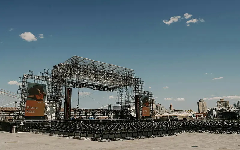 Empty outdoor concert stage with seating and banners showing Diana Ross under a clear blue sky.