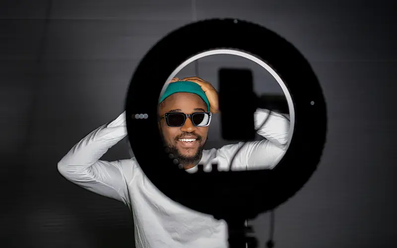 Smiling man wearing sunglasses and a teal beanie posing behind a ring light with a phone holder.