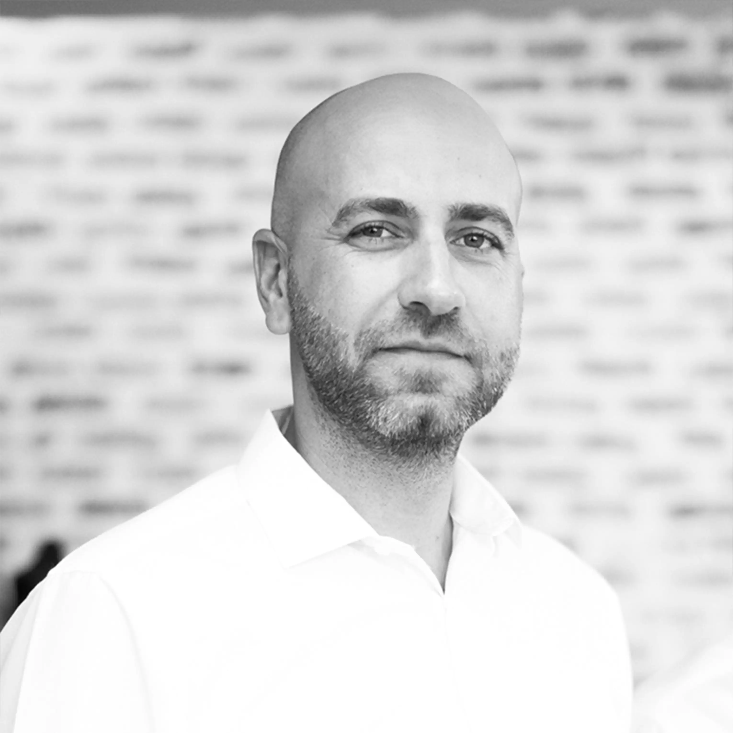 Black and white headshot of a bald man with a beard wearing a white collared shirt, smiling slightly.