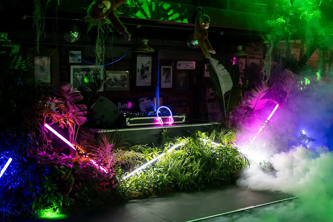 Indoor jungle themed DJ setup with neon lights, plants, fog, and hanging toy monkeys.