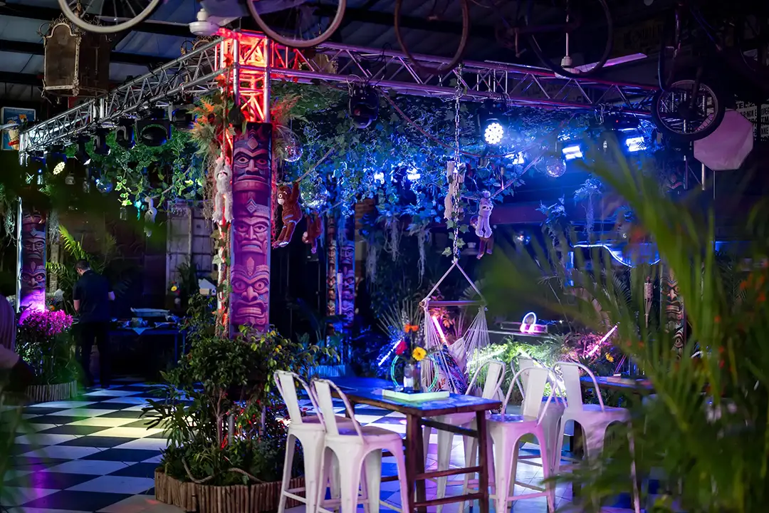 Indoor tropical themed cafe with tiki pillars, hanging plants, white chairs, and mood lighting.