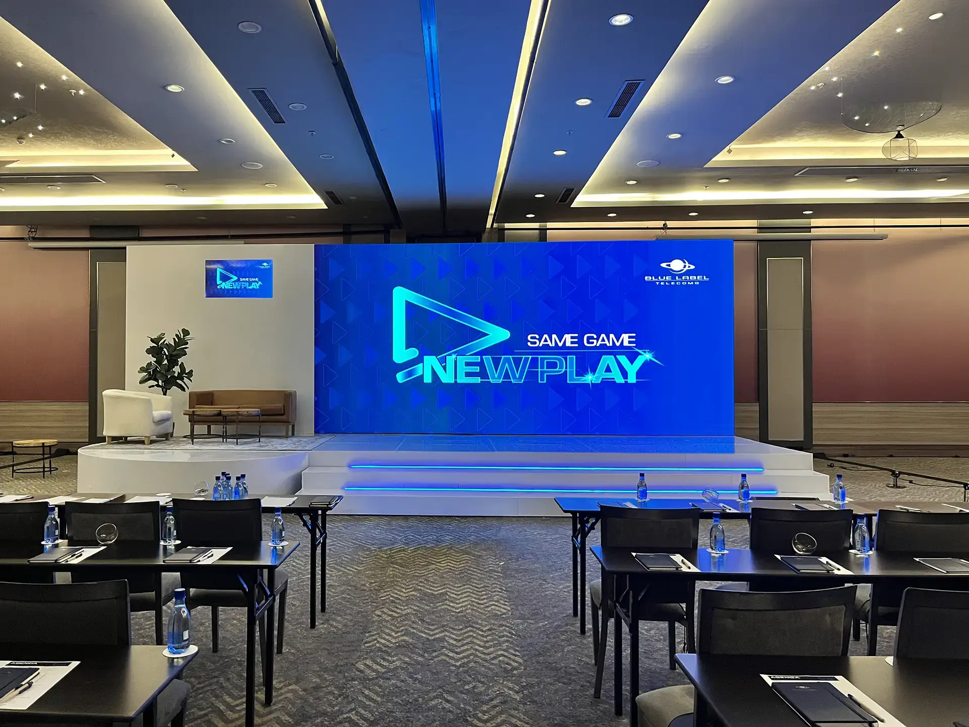 Conference room with stage, blue screen showing SAME GAME NEW PLAY, and rows of chairs with tables.
