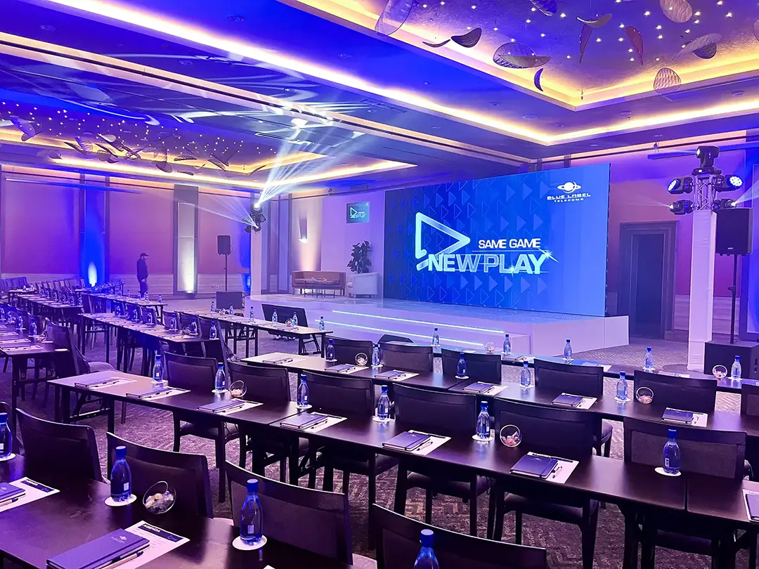 Conference room with rows of tables, chairs, water bottles, and a blue-lit stage with a large screen reading New Play.