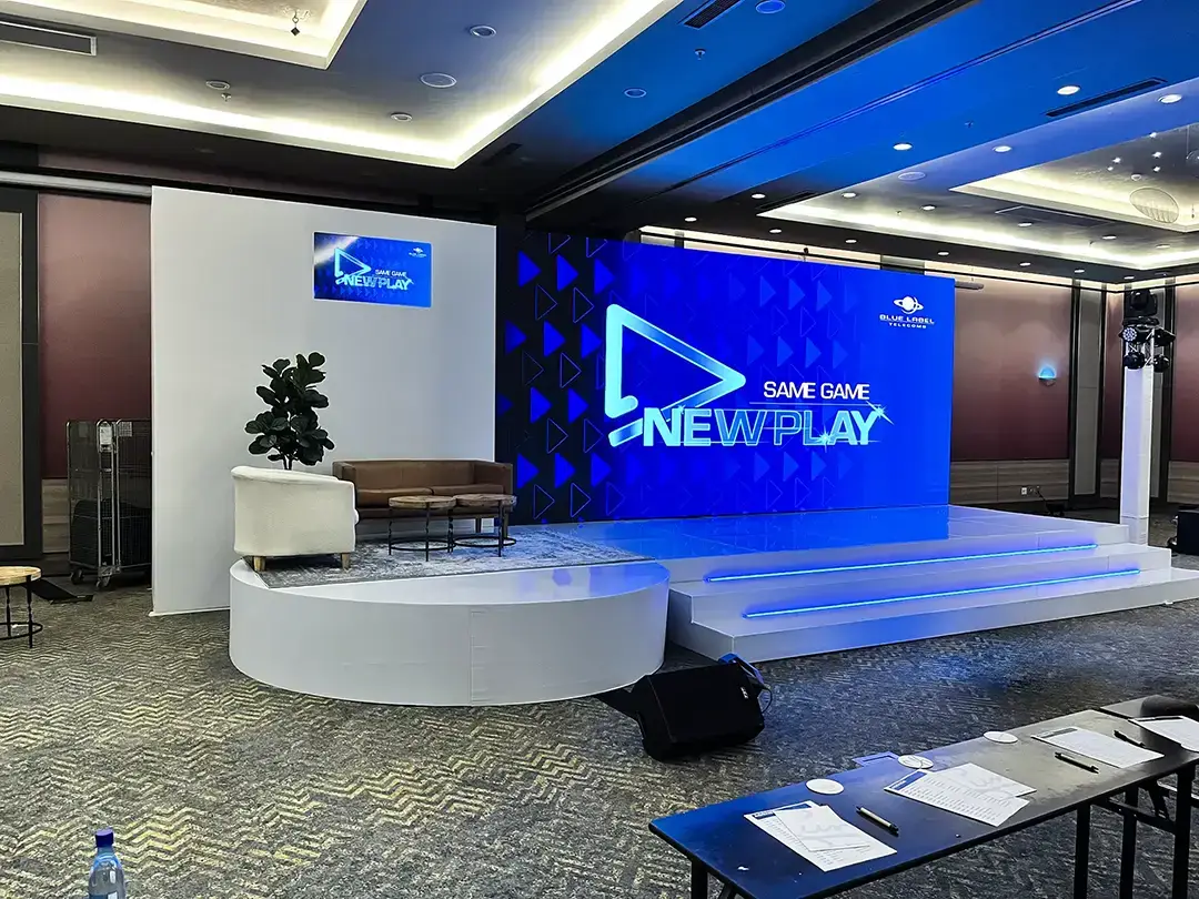 Conference stage with blue digital screen showing Same Game New Play logo and seating area with sofa and chair.