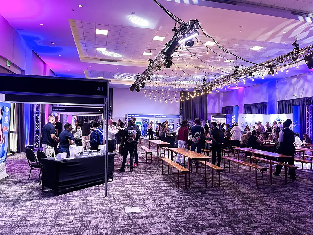 Indoor event with people networking at booths and sitting at wooden benches under string lights.