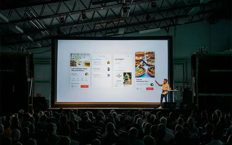 Man giving presentation on stage with large screen showing app interfaces to seated audience.