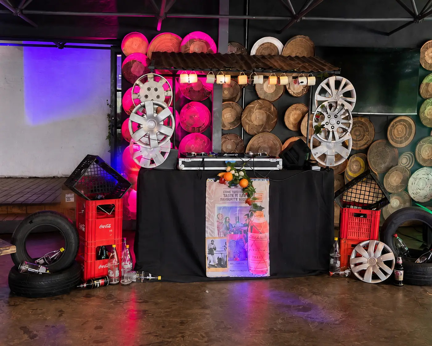 DJ setup with car tires, Coca Cola crates, hubcaps, and woven basket wall decor lit by pink and blue lights.