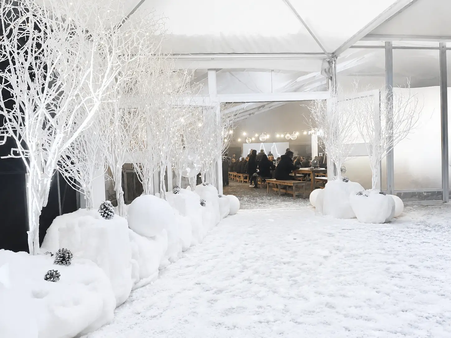 Snow covered path lined with white lit trees leading to a tent with people sitting at wooden benches.