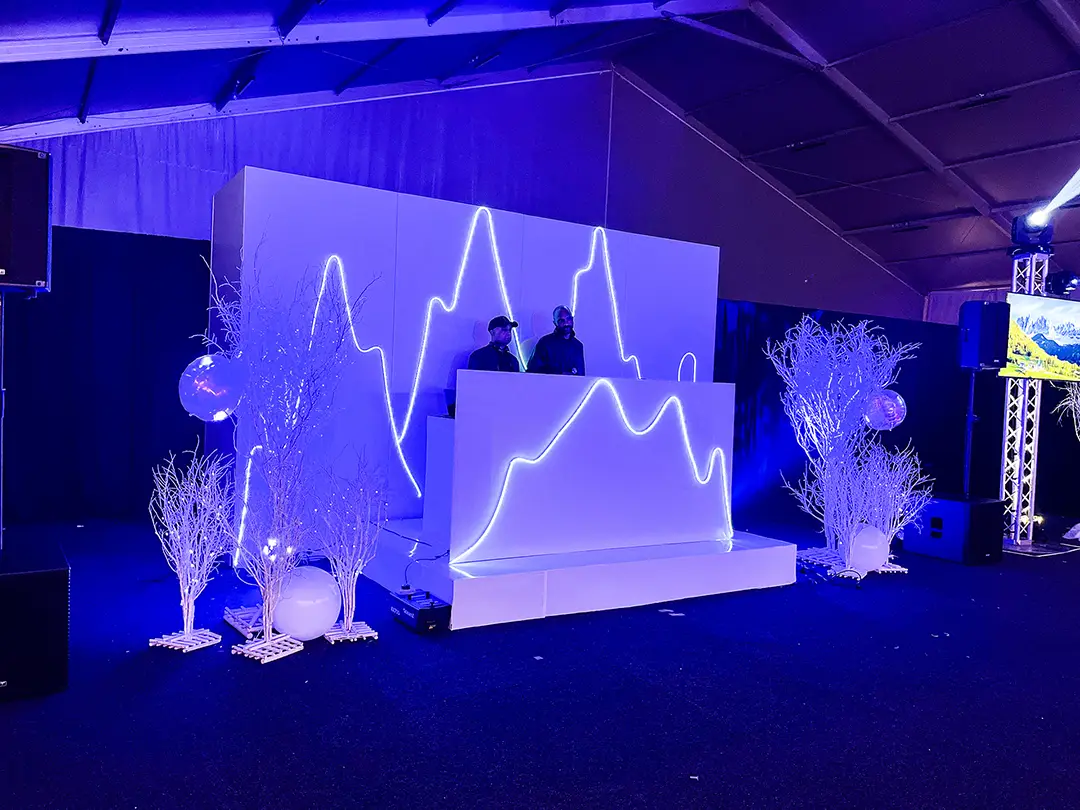 Two DJs performing behind a white booth lit with blue neon mountain-shaped lights in a dark indoor venue.