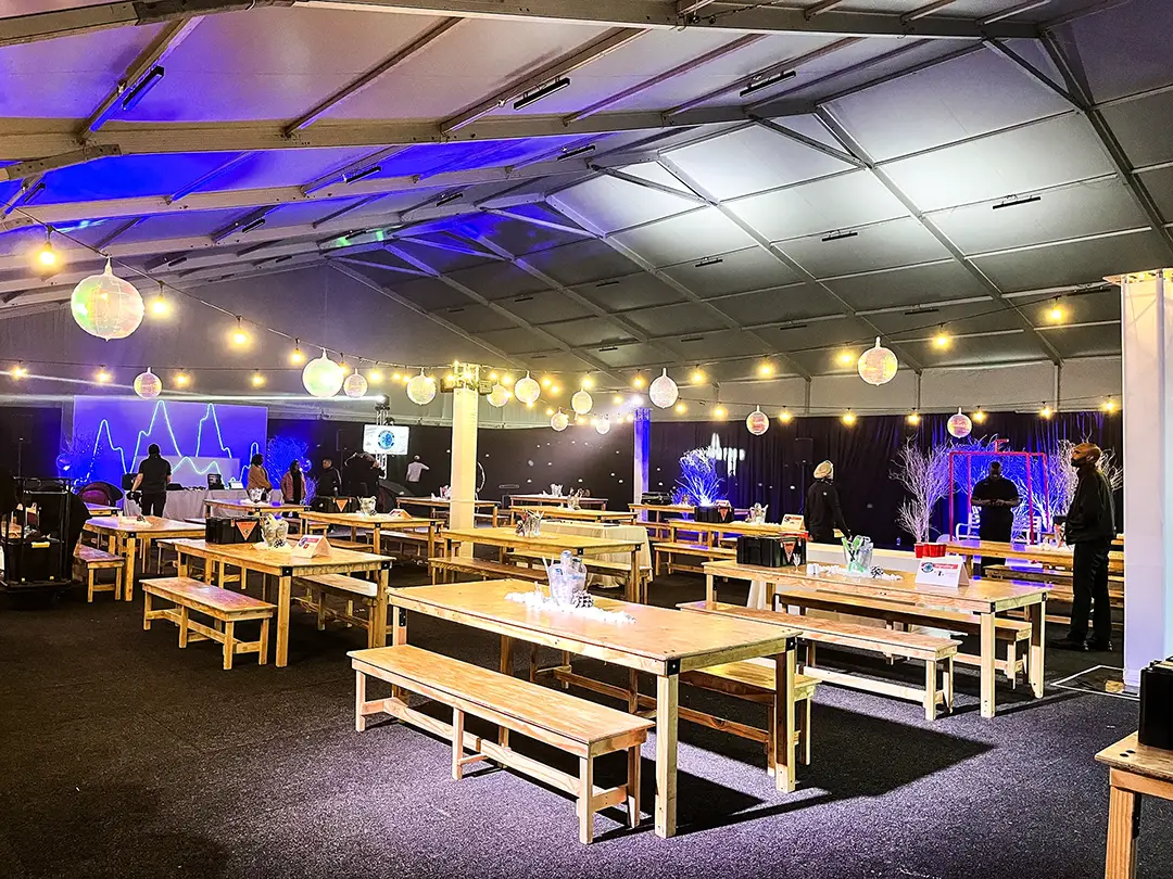 Indoor event space with wooden picnic tables and hanging lantern string lights under a large tent.