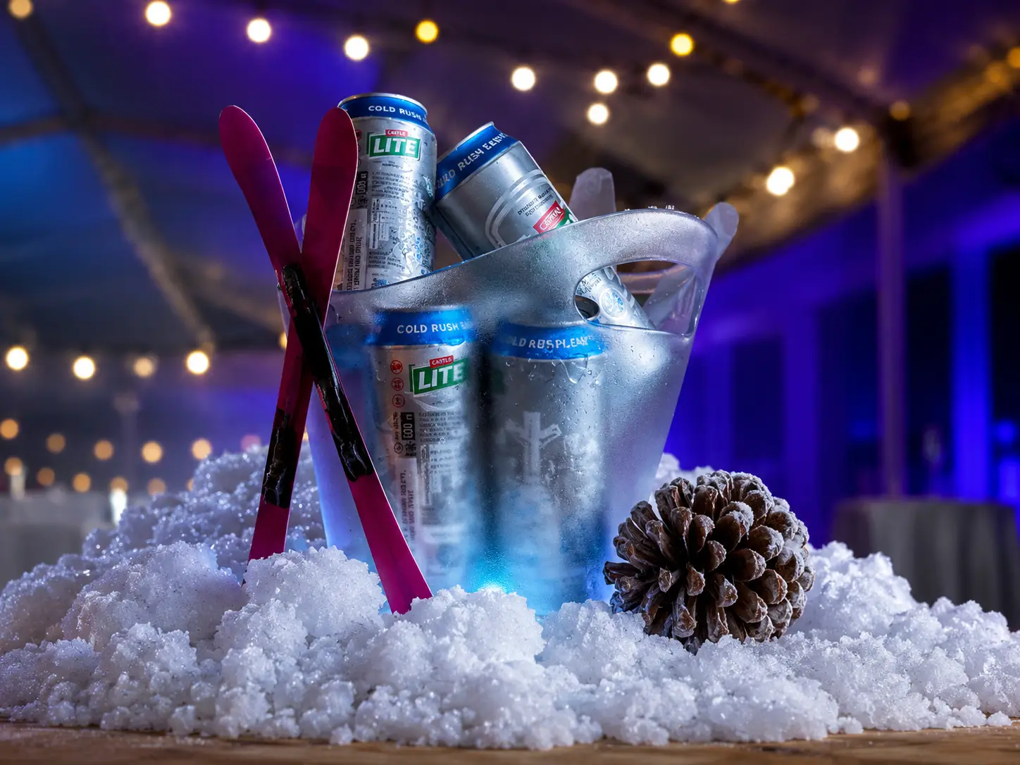 Ice bucket with cold cans of Castile Lite beer, pink ski poles, pine cone, and faux snow on table.