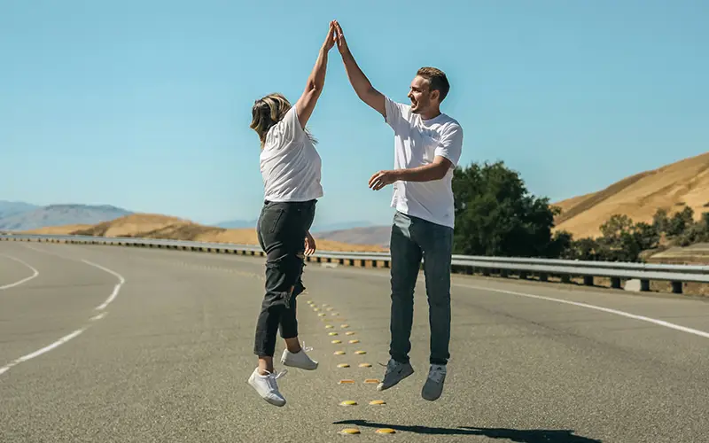 Man and woman jumping and high-fiving each other on a deserted highway with hills in background.