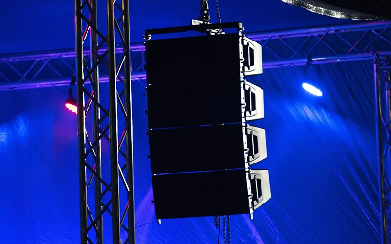Large black speaker array hanging on metal trusses with blue and purple stage lights.