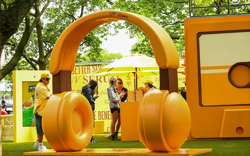 Large yellow headphone sculpture in park with people and yellow promotional displays around it.