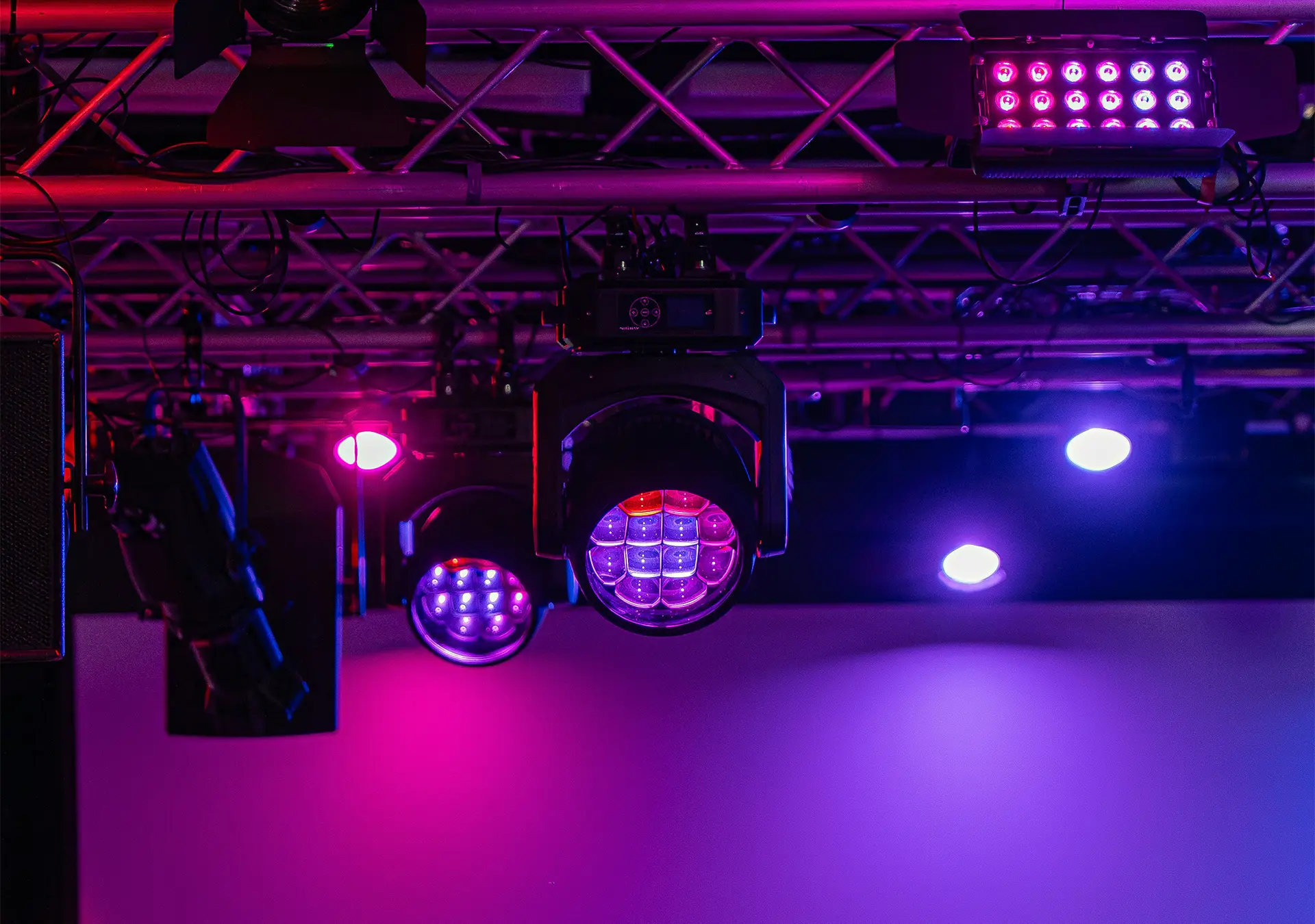 Stage lighting fixtures hanging from a metal truss with purple, pink, and blue lights shining.